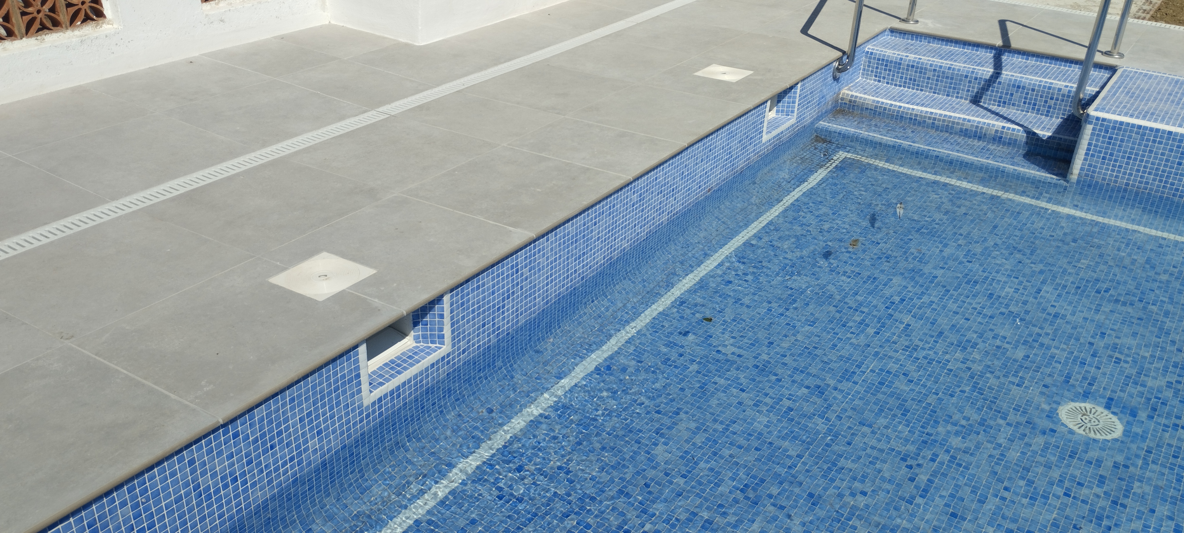 Detalle de las aberturas de los skimmers en la pared de una piscina con mosaico azul y escalera de obra